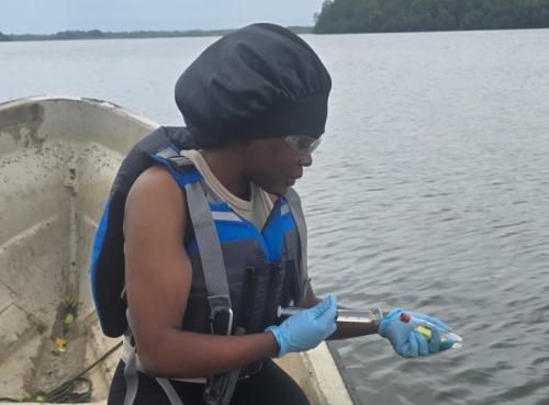 A person wearing a life jacket and gloves holds a sample bottle on a boat near water.